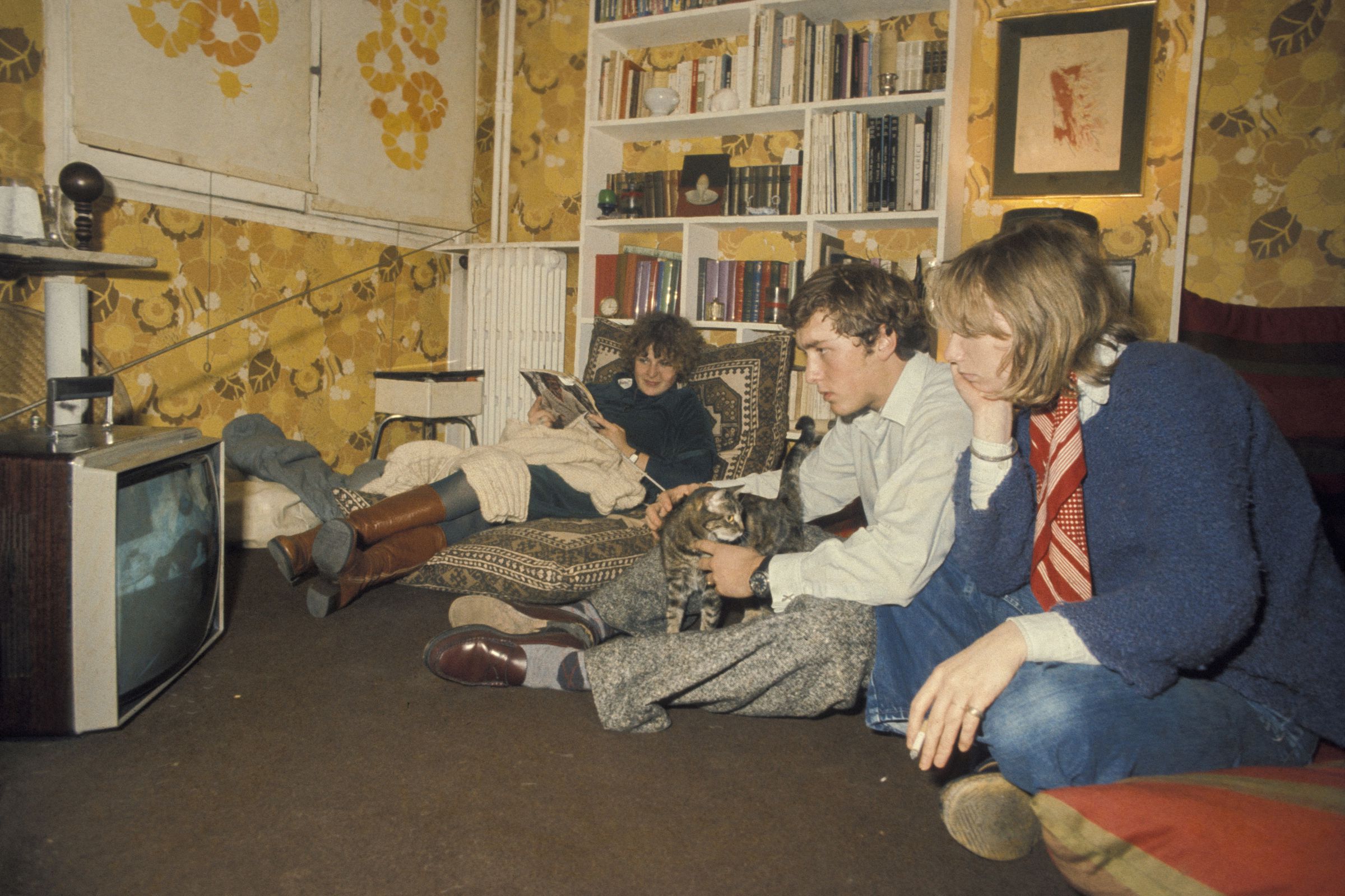 A group of adults sit in a living room in ’70s. They are on the floor watching a TV also on the floor with rapt attention.